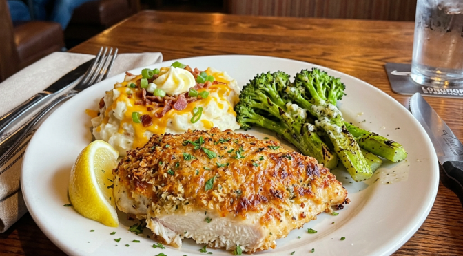 Grilled chicken breast with mashed potatoes topped with cheese, bacon, green onions, and grilled broccoli with a lemon wedge
